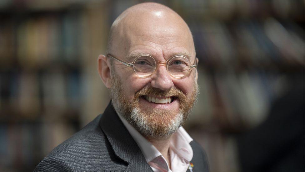 A bald man with glasses smiles at the camera. He has a ginger and grey beard and is wearing a grey suit and pink shirt.