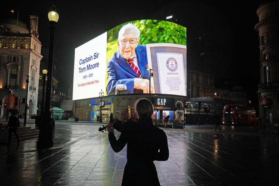 A woman plays the violin in Piccadilly Circus in front of a screen showing a picture of Captain Sir Tom Moore