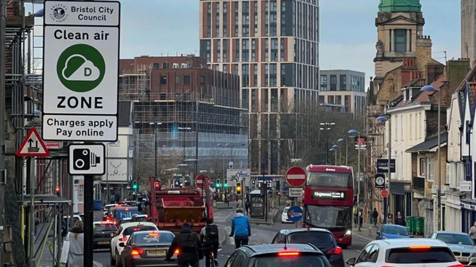 Image of Bristol traffic. Lots of cars, buses and cyclists can be seen in a traffic jam. Buildings can be seen in the distance and a white sign indicating a clean air zone can be seen to the left of the frame