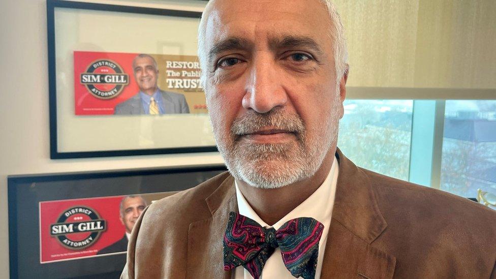 Sim Gill wearing a brown suit jacket and bowtie looking into the camera, in an office with campaign photographs of himself on the wall