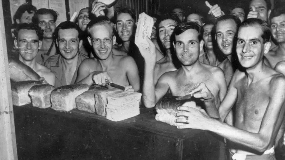 Men, mostly without shirts and looking malnourished, are crowded around loaves of bread on a table. Some have picked up slices of bread to wave in the air.