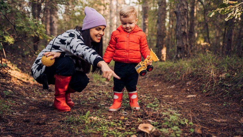 A mother smiles as she points something out to her toddler on a forest walk. They both wear red wellies and warm coats and the little boy holds a yellow digger truck in his hand.