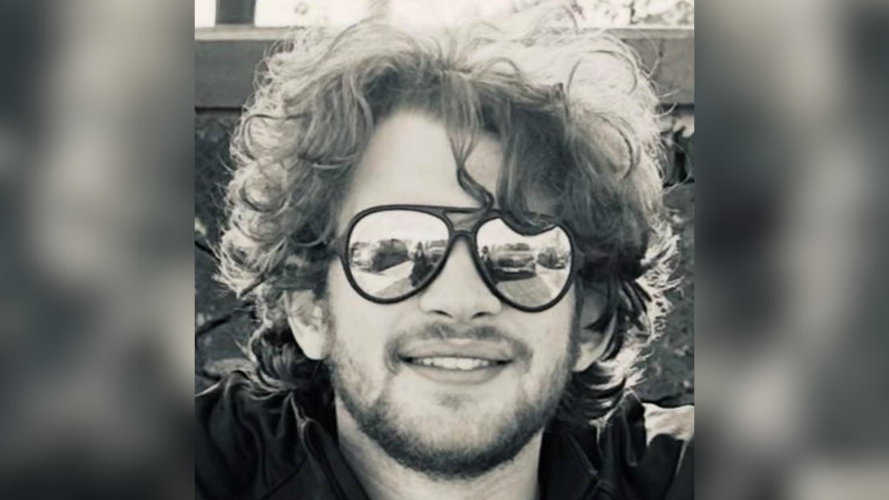 A black and white image of Thomas Chapman. He has long, curly hair and is wearing large sunglasses while smiling.