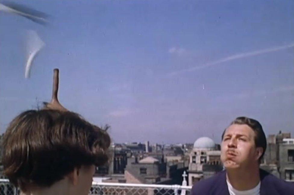 Rondart (aka Ronnie Tomlinson) blowing a dart out of his mouth, through a cigarette perched on a woman's head, into a dart board