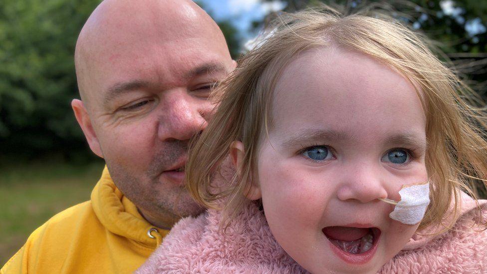 Terry, wearing a yellow hoodie, cuddling his daughter Beatrix who is wearing a pink fluffy coat and has a feeding tube held in place in her nose by white surgical tape. Both are smiling.