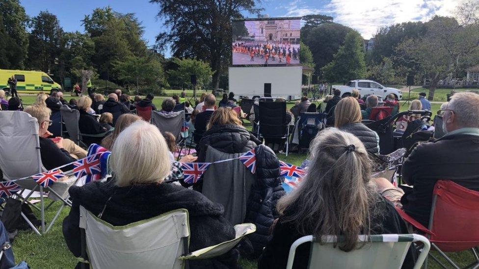 People sitting on the grass or on deck chairs watching a big screen