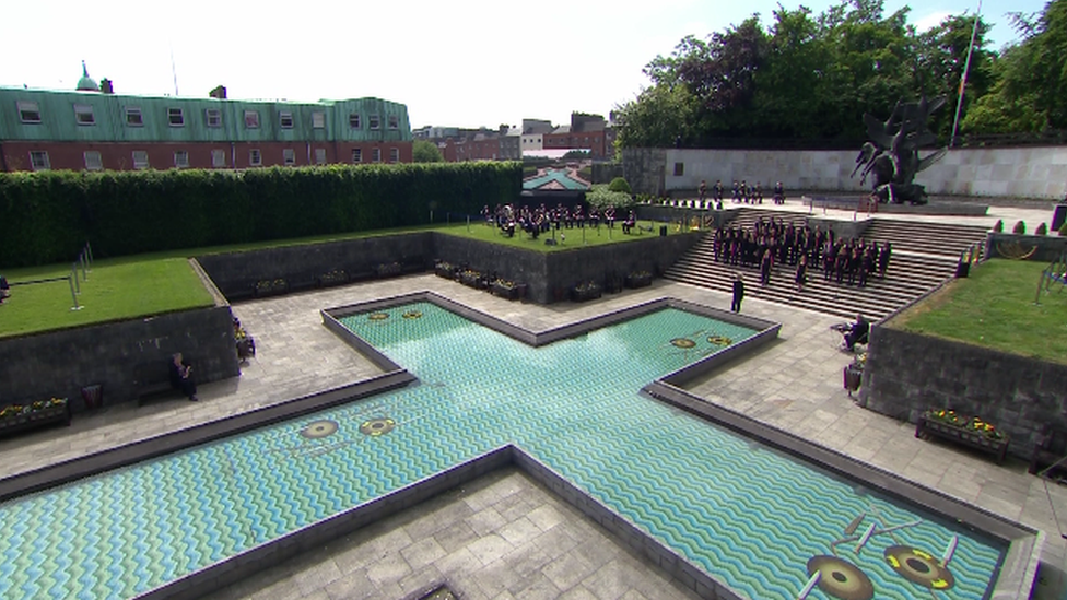 A wide shot of the Garden of Remembrance in Dublin, focussing on the pool of water shaped like a cross and a gathering of people on steps beyond