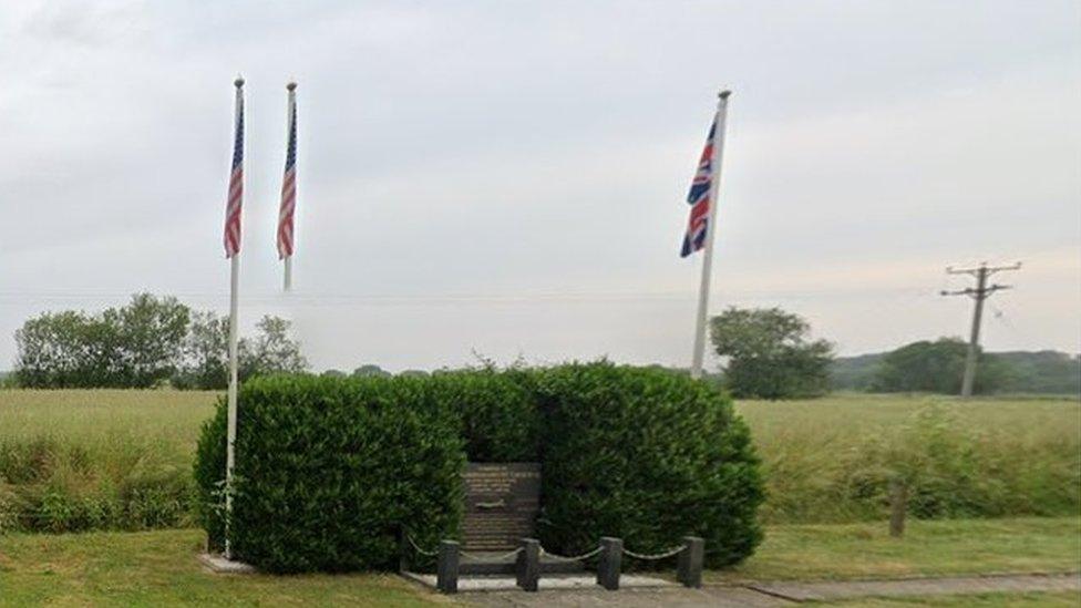 Small inscribed stone surrounded by hedge. There are three flags around the memorial on the edge of an airfield.