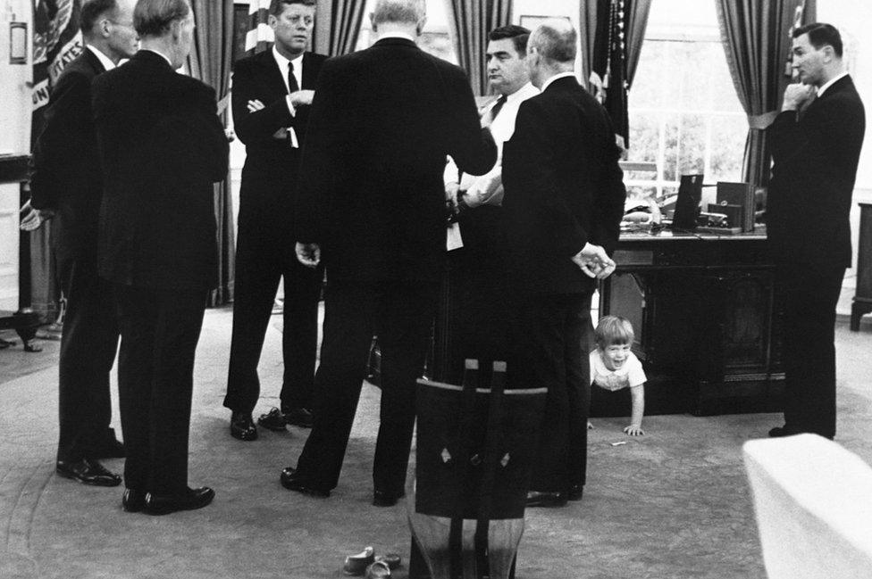 President Kennedy meets with advisors in the Oval Office, while his son, John Jr. plays under the desk. 1963