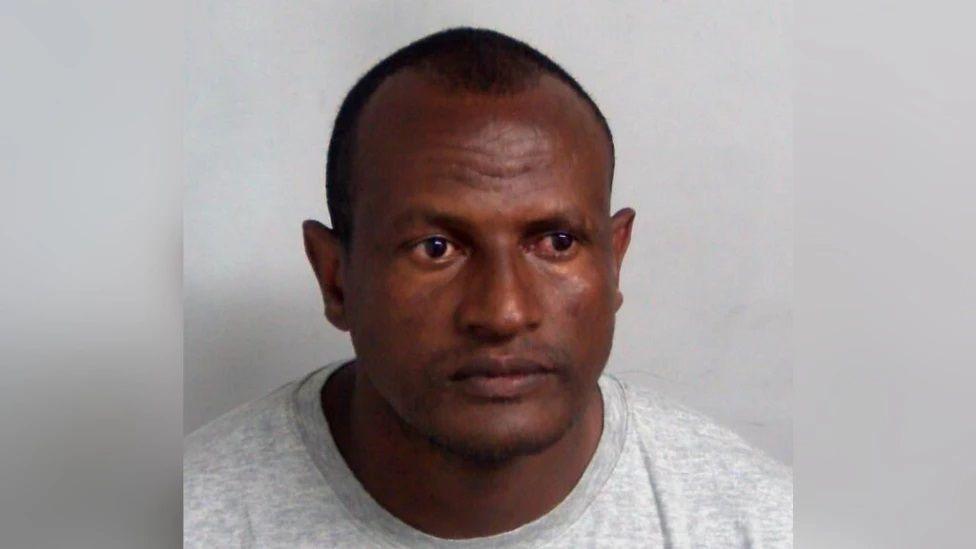 Hadush Kebatu with very short dark hair wearing a grey T-shirt against a plain white background in a police custody photo