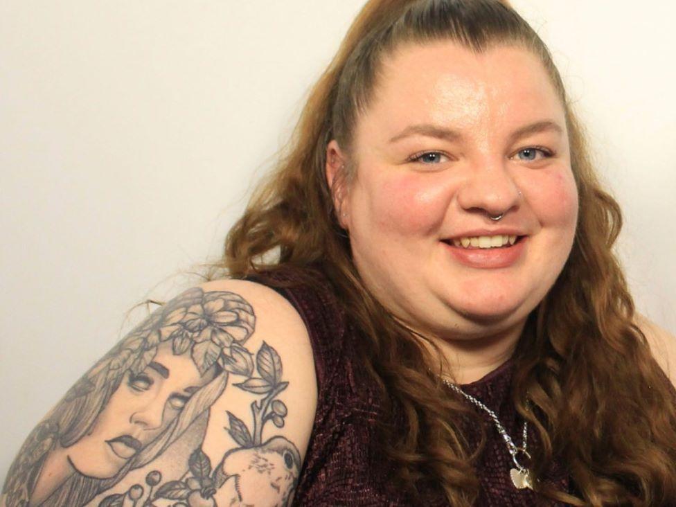 A woman with wavy brown hair and tattoos on her arm smiles at the camera