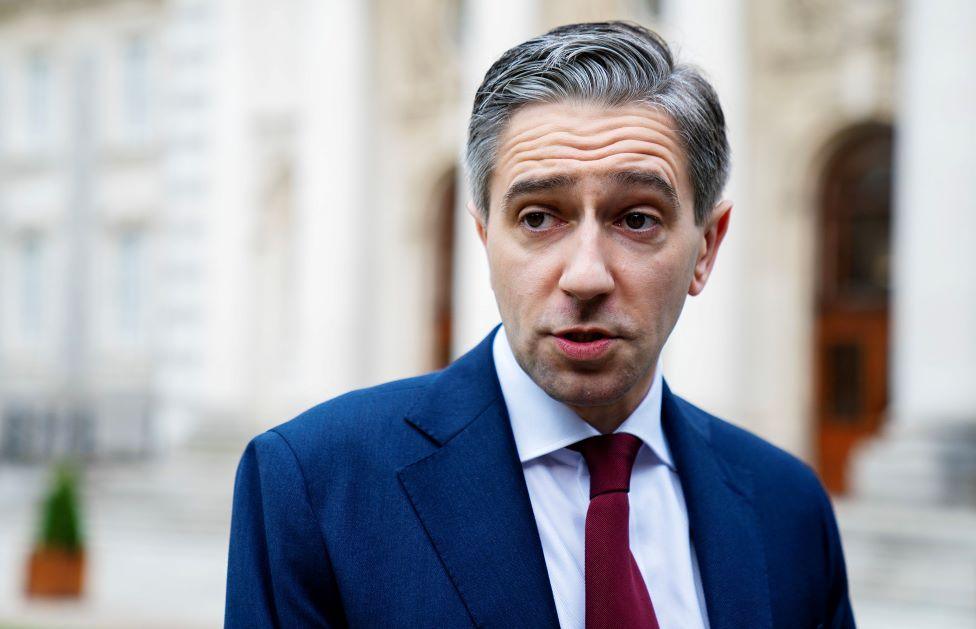 Taoiseach Simon Harris stands in a blue suit with a red tie in front of a blurred out background