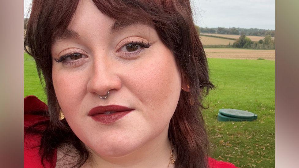 Frankie, a young woman with brown hair and brown eyes. She is wearing red-brown lipstick and light eye makeup. She is standing in a grassy area with fields in the background