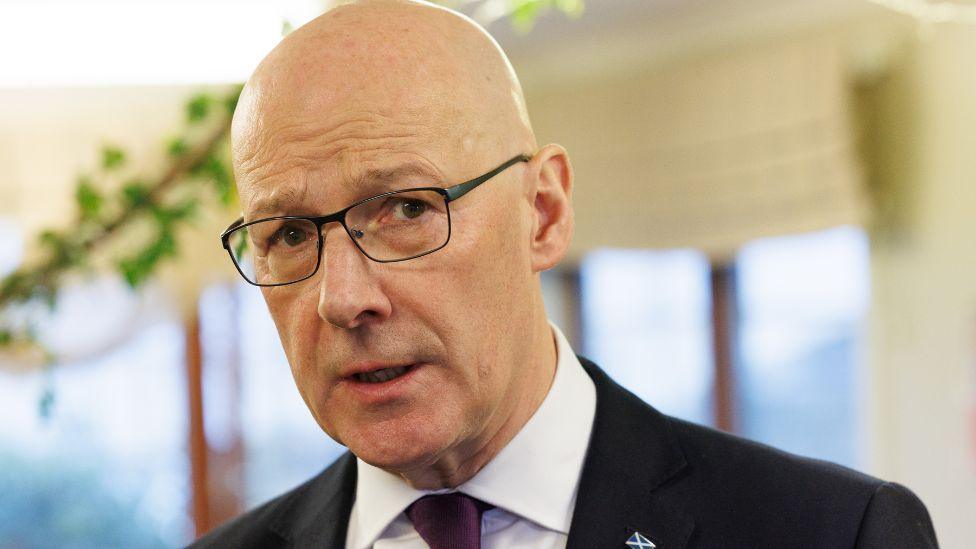 John Swinney, a bald man with glasses wearing a darks suit and white shirt, looks towards the camera in a close-up shot 