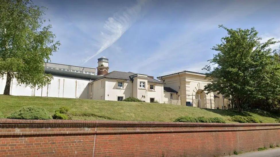 A screenshot from Google street view of the outside of the prison. It's a white building behind a grassy hill and a brick wall.