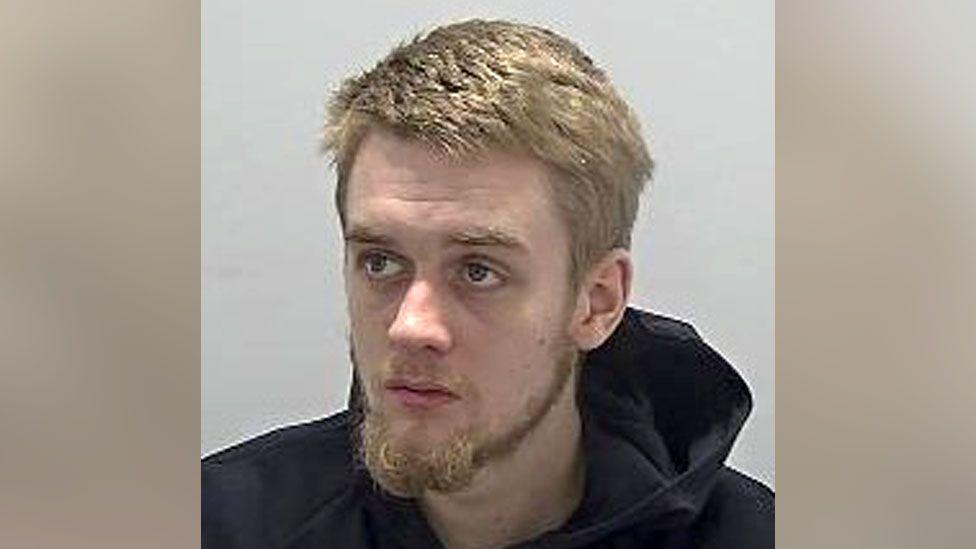 A mug shot of a man with light brown hair wearing a black hooded top. He is looking away from the camera and has a short moustache and beard.