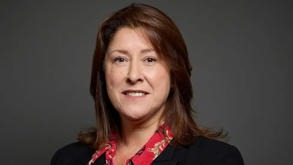 A portrait of Michelle Scrogham who has brown, straight hair. She's smiling at the camera and is wearing a red patterned shirt and black jacket.