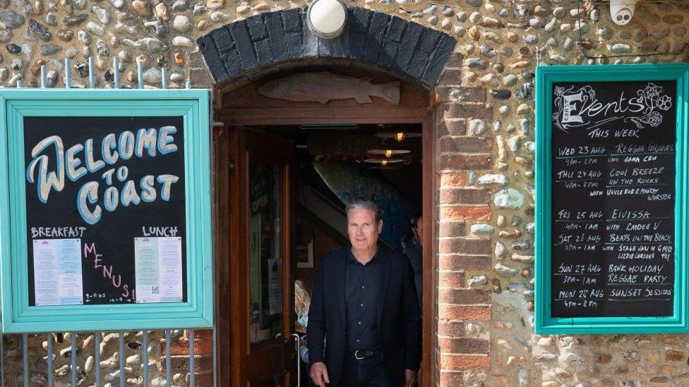 Labour leader Sir Keir Starmer visits Worthing in West Sussex. He is standing in the doorway of a cafe looking at the camera and wearing a dark blue shirt and suit jacket. Either side of him on the brick wall of the front of the cafe are black chalkboard signs advertising the breakfast and lunch menus to the left and then the events for the week to the right.