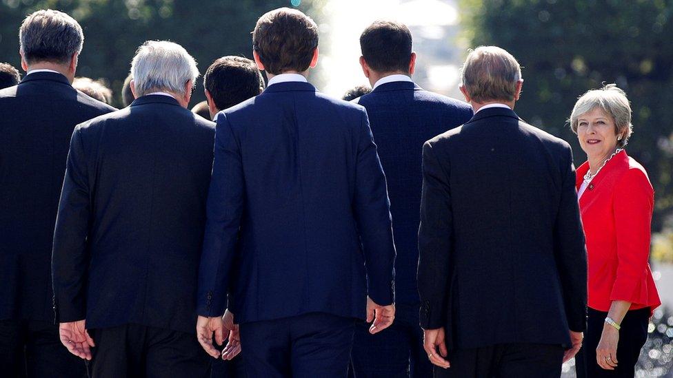 Theresa May, in red, faces the camera while EU leaders in dark suits walk away from the camera.