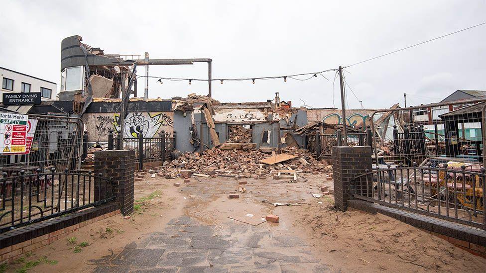 View of the demolished building. Part of the curved grey wall to the left is standing, the wall below it has graffiti on and there is a pile of rubble to the front.