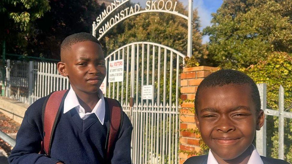 Brothers Anele (left), who is carrying a rucksack, and Lifalethu Mbasana in their navy jumper and white shirt uniforms outside their school gate in Simon's Town. Incorporated into the the white-painted metal gate is a curved sign reading 'Simonstad Skool'