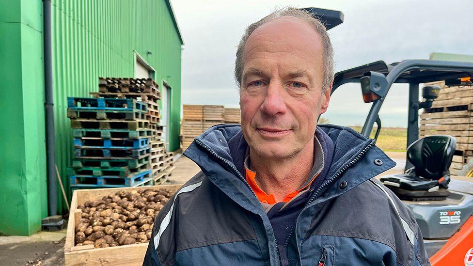 Farmer David Armstrong wearing a blue rain jacket stands in front of a fork-lift truck carrying a box of potatoes 