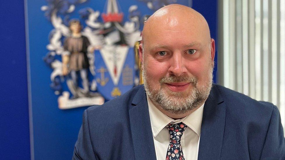 Man with grey beard wearing suit and tie and sitting in front of a council crest