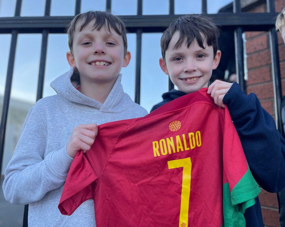 Andrew and Ciaron hold a red Ronaldo top in front of the away gate 