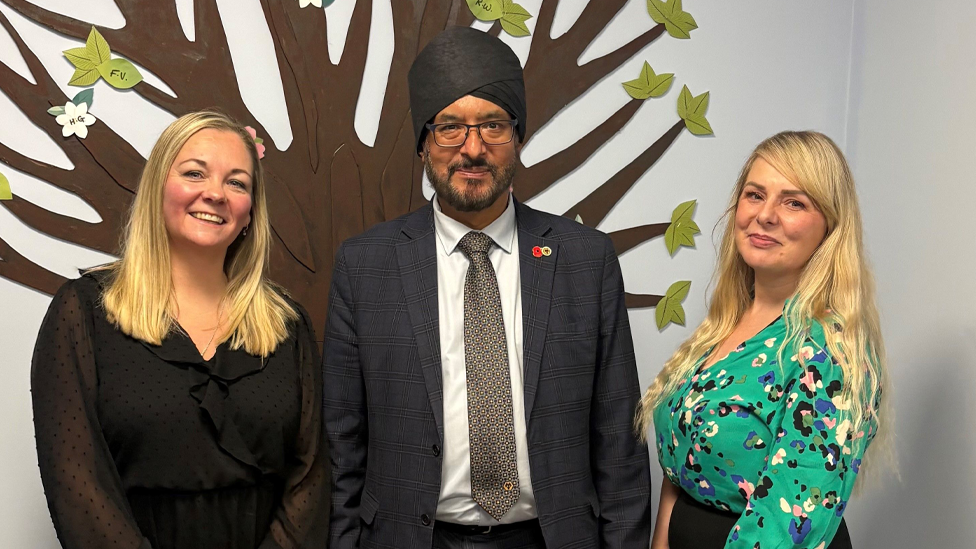 Emma Rennie-Gibbons and Samantha Webster stand either side of MP Warrinder Juss. In the background is a tree painted on to the wall of the school