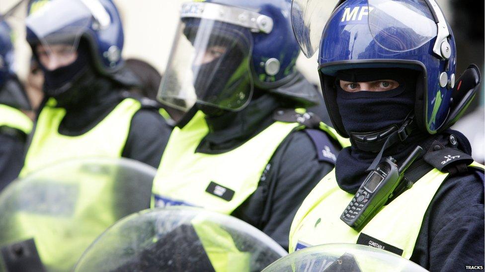 A row of three police officers dressed up in riot gear, which includes a helmet and scarf covering all but their eyes. One of the officers is looking directly into the camera.