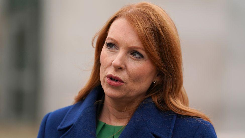 Ash Regan, a woman with red hair, looks to her right while speaking. She is visible from the shoulders up and wearing a blue jacket and green top. 