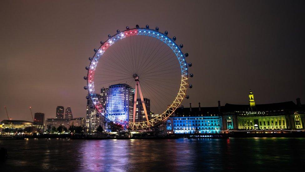 The London Eye, half lit up in red, white and blue and half in yellow and blue, next to a yellow and blue City Hall.