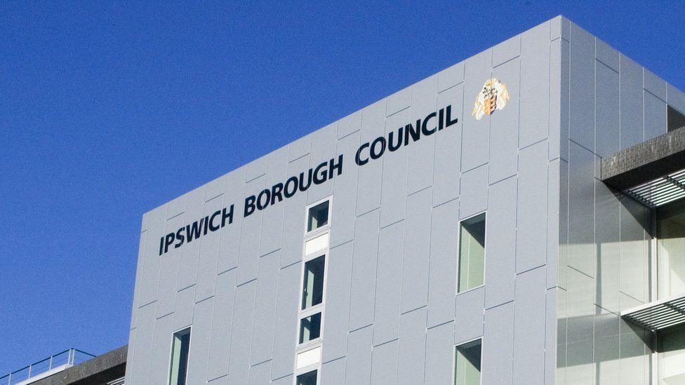 A view of the top of the Ipswich Borough Council building. The grey building has the name of the council written on it in large black letters. Small windows can be seen on the building.