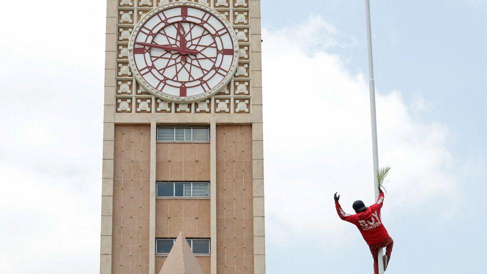 A mourner of Raila Odinga dress in red and carrying a palm frond climbs a pole at Kenya's parliament in front of the clock tower.