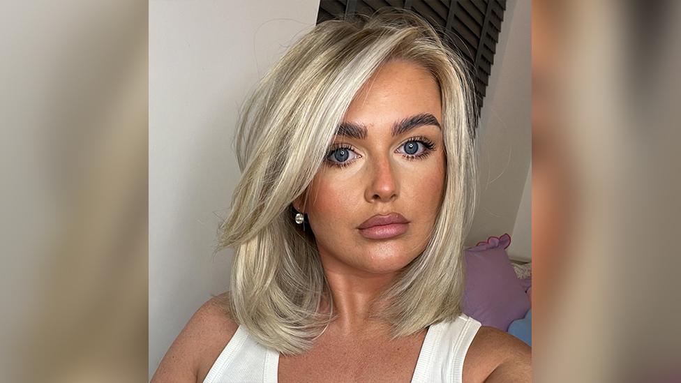 Tamzin McGrath taking a selfie. She is a white woman with short blonde hair and has her make-up done. She is wearing a white vest and there is a pink cushion and dark blinds visible in the white walled room behind her