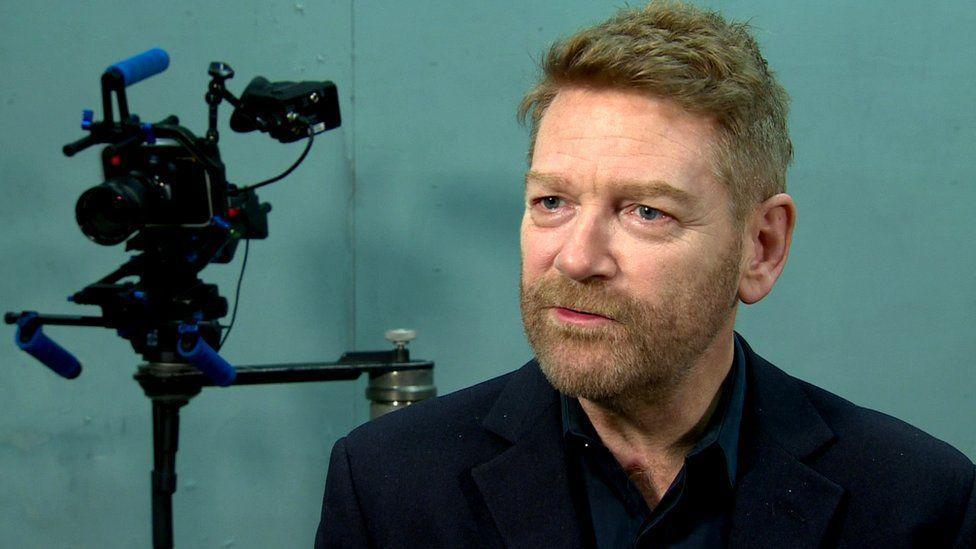 Sir Kenneth Branagh wearing a black shirt open at the collar. He has a short light-brown full beard and short hair.