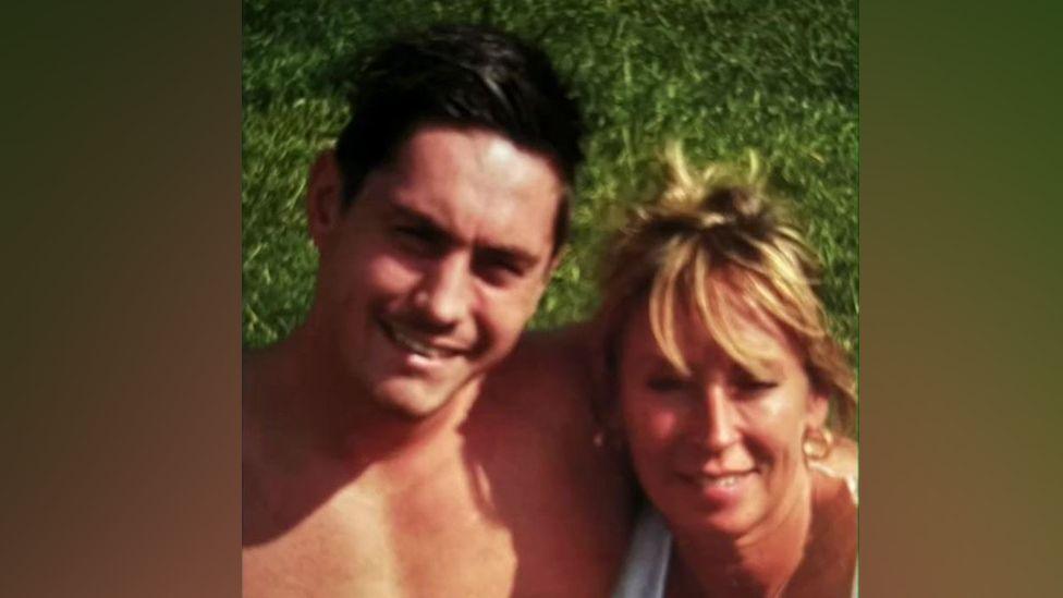 Jamie Attwood with short dark hair smiling at the camera with his arm around his mother, who has medium-length brown hair and is wearing a white top.  They are outside standing on grass.