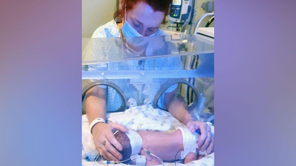 Picture of a woman wearing a face mask and a blue and white hospital gown leaning over an incubator which has a small baby inside. She has one hand on the baby's head and another rests on its nappy. The baby has wires attached to his body and is lying his front on a white patterned blanket. 