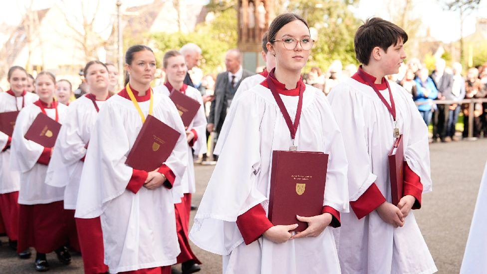 Koristët e katedrales duke mbërritur në Katedralen e Shën Asafit. Ata kanë veshur rroba të bardha me një veshje të kuqe me mëngë të gjata poshtë. Secili prej tyre mban nga një libër të kuq ndërsa ecin në çift.