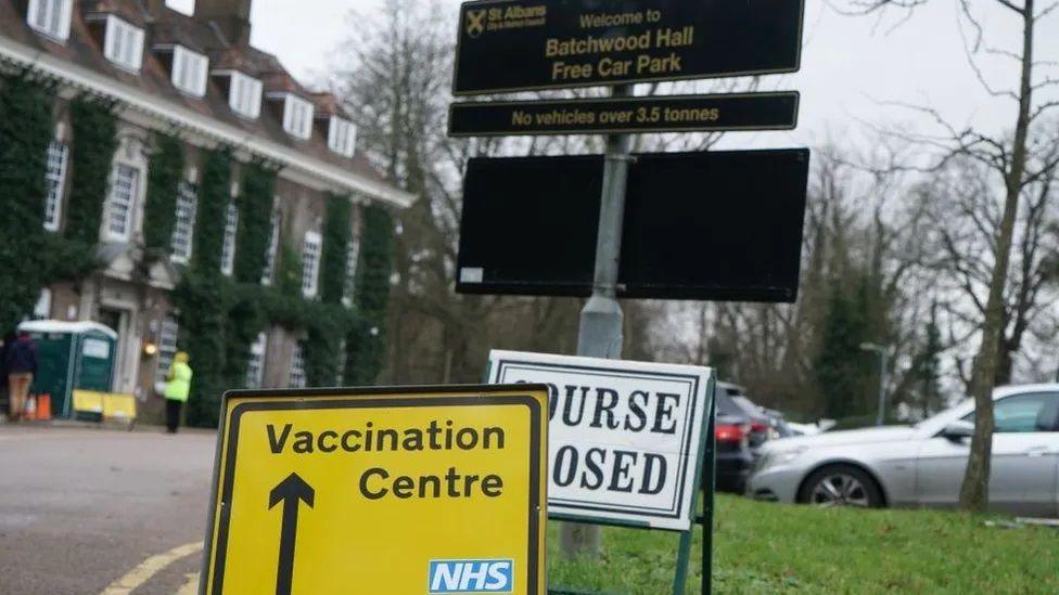 A yellow NHS sign with the words "vaccination centre" in the foreground and a "Batchwood Hall" sign behind it. In the background is an ivy-covered manor house