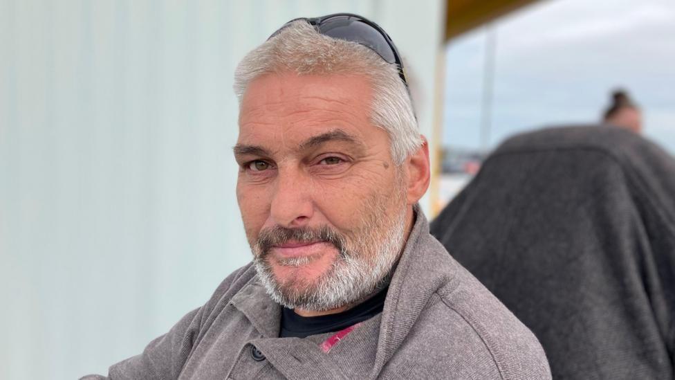 Christopher duke parë kamerën. Ai ka mjekër dhe ka veshur një bluzë gri.