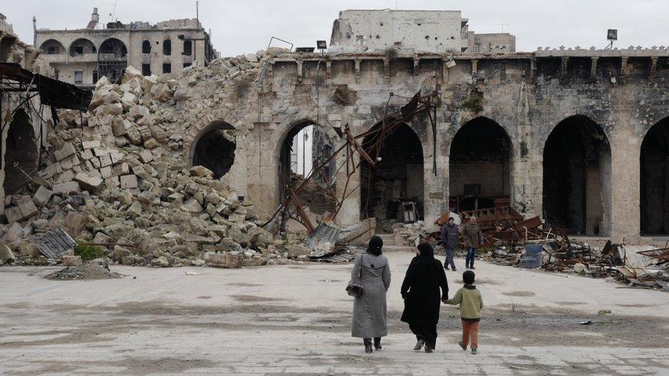 Groups of people walk in front of destroyed buildings