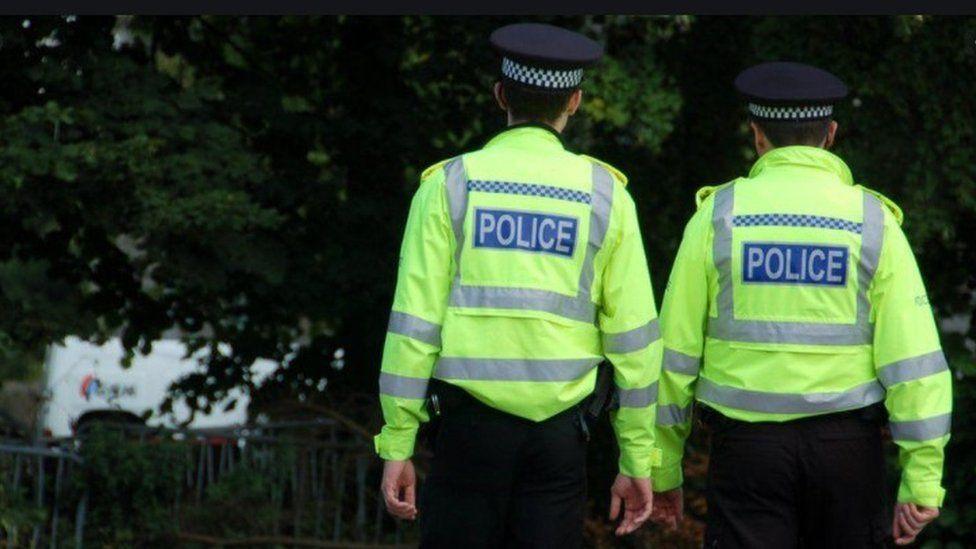 Two police Scotland officers viewed from behind as they look into a wooded area.