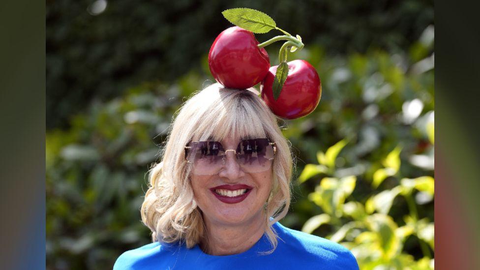 A woman wearing a fascinator which is two large plastic cherries. She is also wearing sunglasses and a blue dress.
