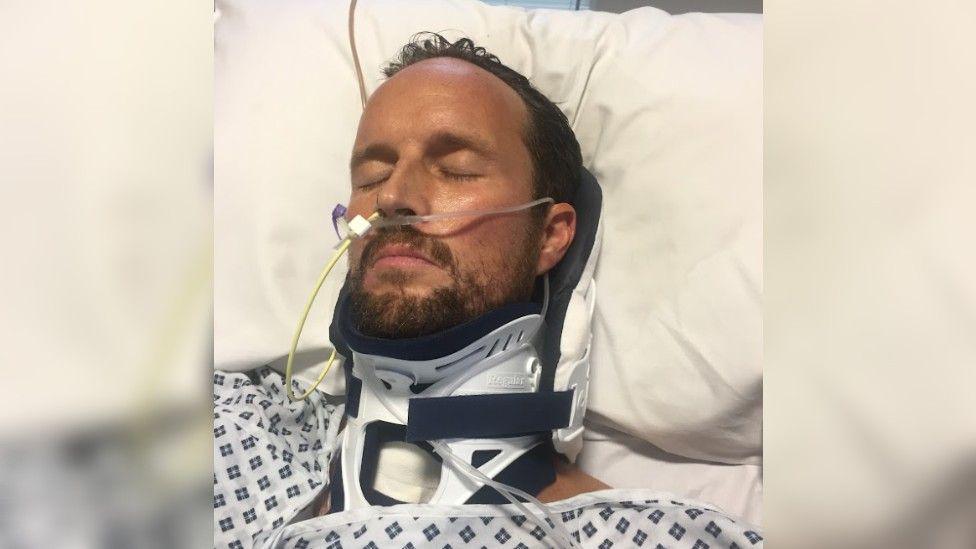 Jamie Rudd in a hospital bed with a neck brace and tubes coming out of his nose