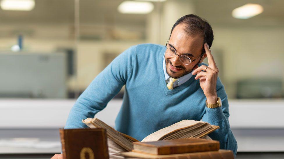 Dr Majid Daneshgar, with a close trimmed dark beard and short dark hair, wearing a light blue v-necked jumper, leaning on one elbow, propping his head up with his fingers and smiling down at a pile of old books