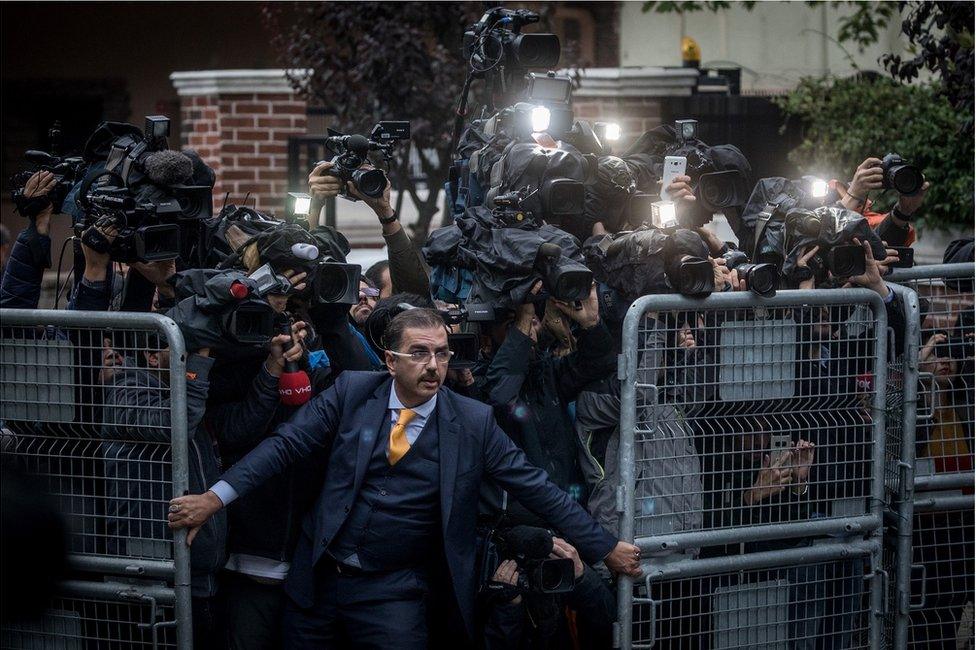 A crowd of cameramen try to get through a gate as a man holds them back