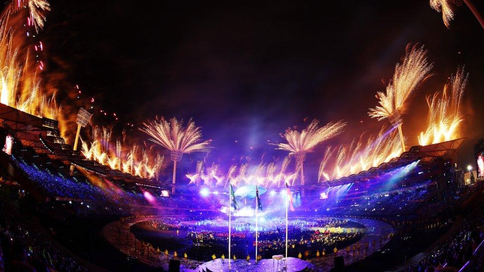 bright fireworks burst from the roof of a large stadium filled with a massive crowd of spectators and athletes