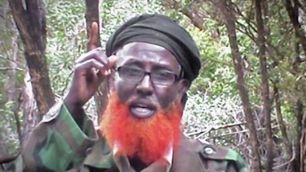 Abdulqadir Mumin, with an orange beard and wearing reading glasses, fatigues and a black headscarf, hold ups his finger to the sky as he talks. Trees and bush can be seen behind him.