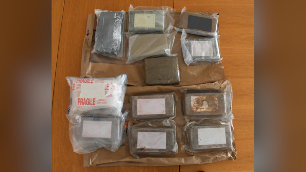 A number of different size bricks of cocaine in plastic bags are laid on top of two brown paper bags on the ground.